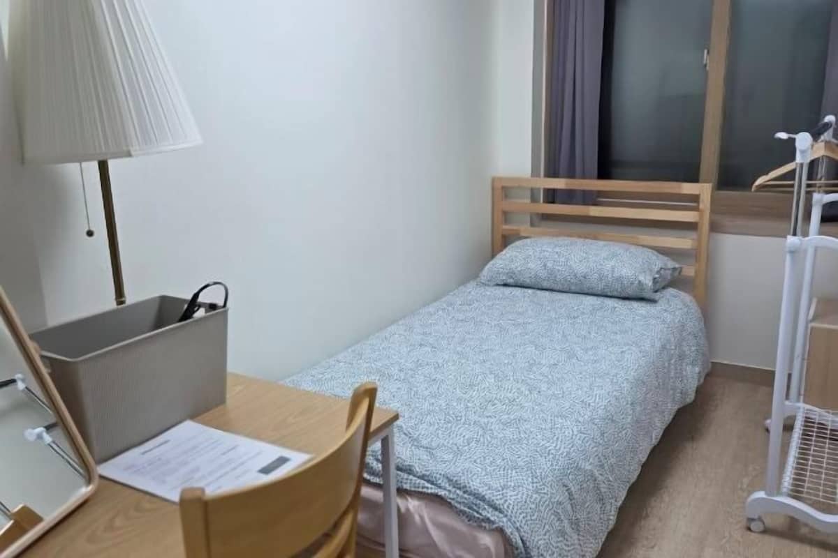 A small, single-bed room with a wooden headboard, a desk with a chair, a lamp, a bedside table, and a window with curtains.
