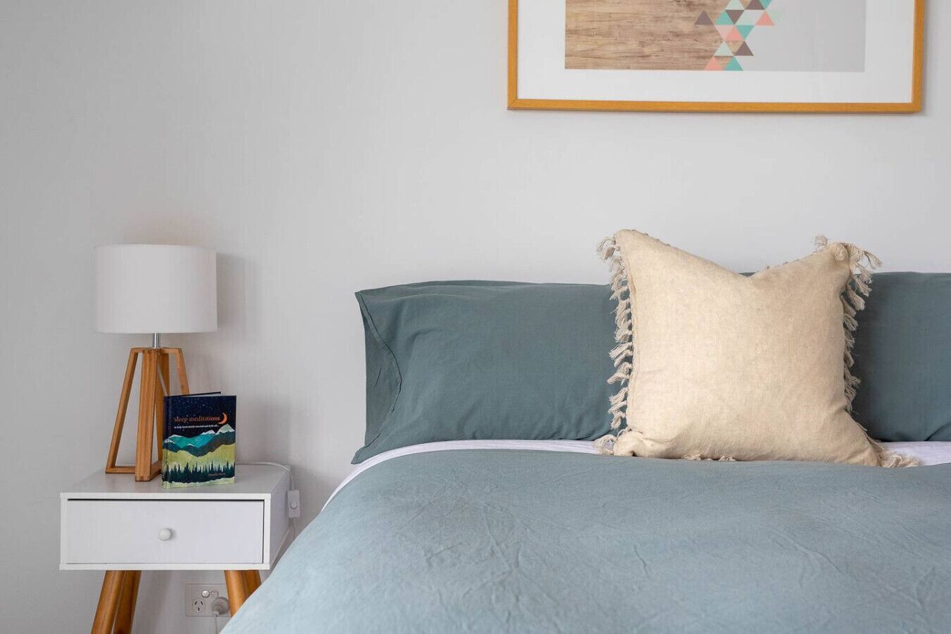 Une chambre à coucher bien rangée, avec un lit, une table de chevet, une lampe et un tableau encadré au mur.