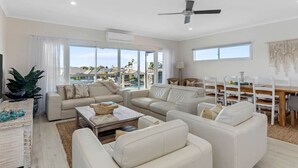 Amplia sala de estar con un gran ventanal, ventilador de techo y un comedor con mobiliario blanco.
