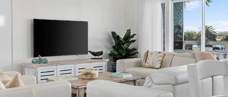 A modern living room with a flat-screen TV, white furniture, a large plant, and a view of palm trees through the window.