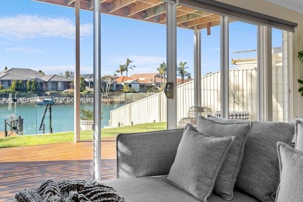 Un salon moderne avec un canapé gris, une terrasse en bois et une vue sur un lac, avec des maisons en arrière-plan.