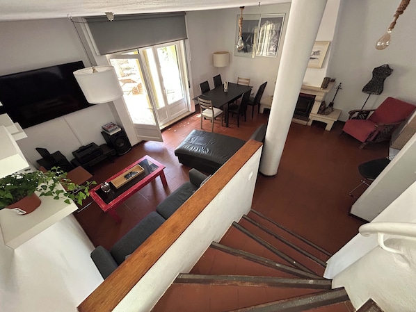 Un vaste espace de vie comprenant un canapé, une table basse et un téléviseur fixé au mur. Un escalier mène à un autre niveau, et un coin repas est aménagé avec des chaises et une table.