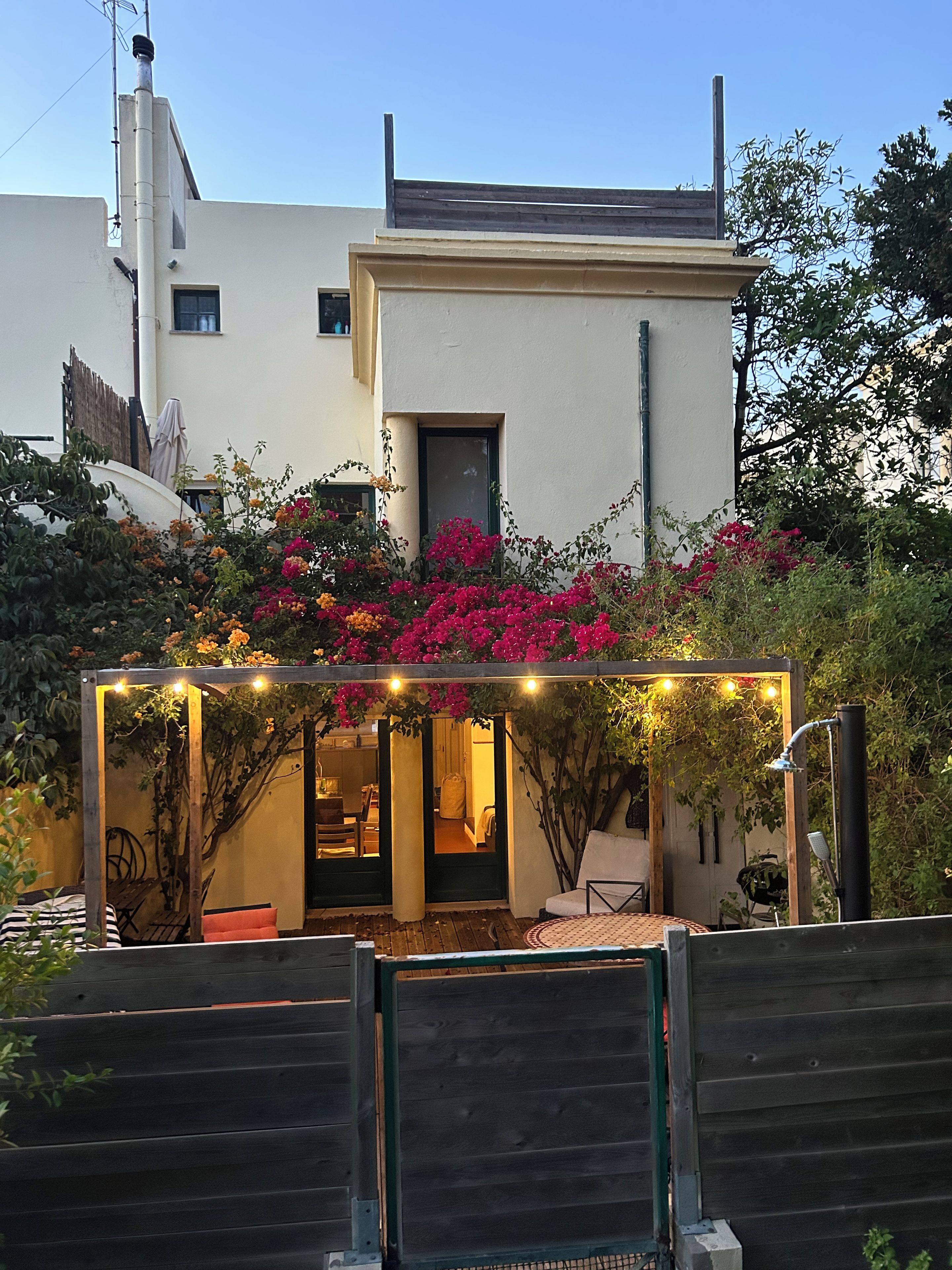 Een modern huis met een overdekte zithoek, omringd door weelderige begroeiing en kleurrijke bloemen.