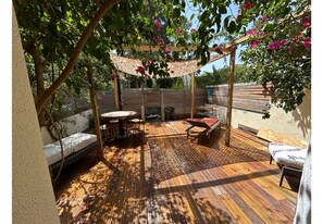 Eine Terrasse im Freien mit Holzboden, einer Pergola mit rosa Blüten und Sitzbereichen, darunter eine Couch und Stühle.