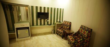 Una habitación con una televisión montada en la pared, dos sillas con estampado, una mesita, un escritorio con espejo y un cubo de basura.