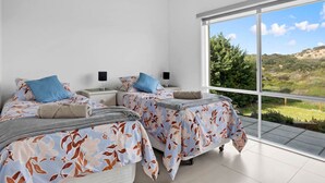 Et moderne soveværelse med to senge, begge med blomstret sengetøj og blå puder, et stort vindue med udsigt over grøn natur og bakker, samt et rent, hvidt interiør.