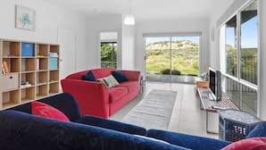 En moderne stue med en rød sofa, en blå sofa og en bogreol i træ. Der er et stort vindue med udsigt over et grønt landskab.