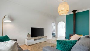 A modern living room with a flat-screen TV on a white console, grey sofas, teal cushions, a woven light fixture, and a visible fireplace. - Berechiah Cottage—Stroll to Cafés & Coast (Port Elliot)