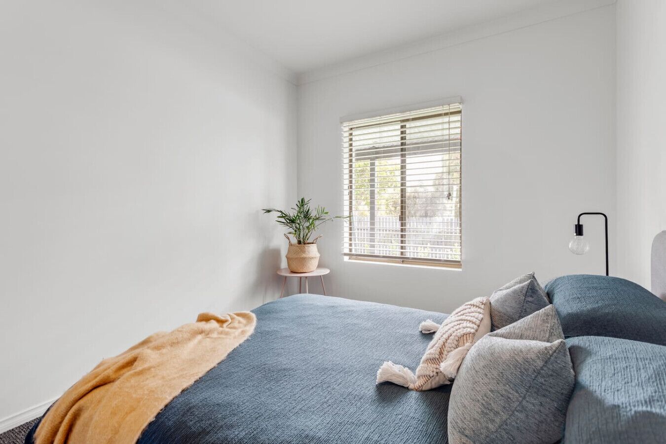 A bedroom with a bed, a bedside table with a plant, a window with blinds, and a floor lamp.