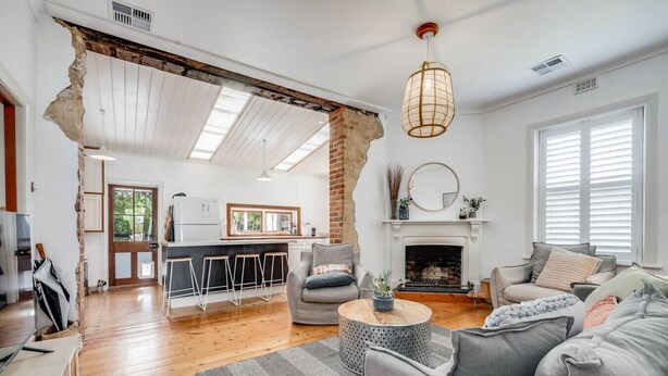 A spacious living room with a fireplace, a kitchen in the background, and a large window with blinds. - Red Door Cottage - Heritage Warmth by the Sea (Port Elliot)