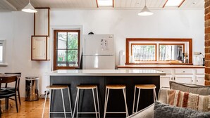 A modern kitchen with a central island, bar stools, a refrigerator, and a window with a view of greenery. - Red Door Cottage - Heritage Warmth by the Sea (Port Elliot)