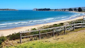 Une plage bordée d’une clôture en bois, avec une eau d’un bleu limpide et, au loin, une côte parsemée de bâtiments et d’arbres.