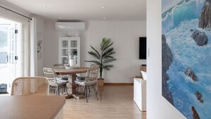 Un coin repas avec une table en bois et des chaises en osier, une plante en pot et un tableau représentant un paysage marin accroché au mur.