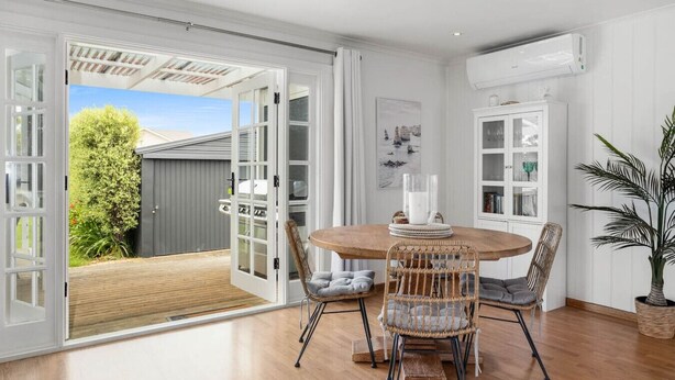 Un coin repas avec une table ronde en bois, des chaises en osier et une vue sur un jardin, grâce à des portes ouvertes.