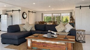 A modern living room with a sectional sofa, a wooden coffee table, and a dining area visible through large windows.