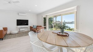 A modern living room with a wooden dining table, white chairs, a leather sofa, and a TV.