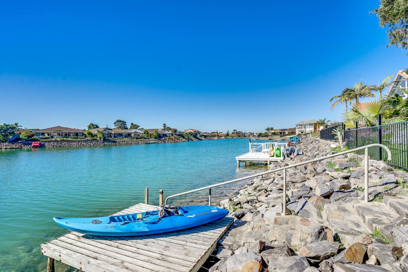 A clear waterfront view with a kayak docked on a wooden pier, a stone walkway, and a row of houses in the background.