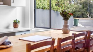 Un comedor con una mesa de madera, sillas, un florero con flores y una ventana con vista a árboles y una valla.