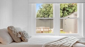 Un dormitorio con un ventanal grande, una cama con ropa de cama blanca y vista a un patio trasero.