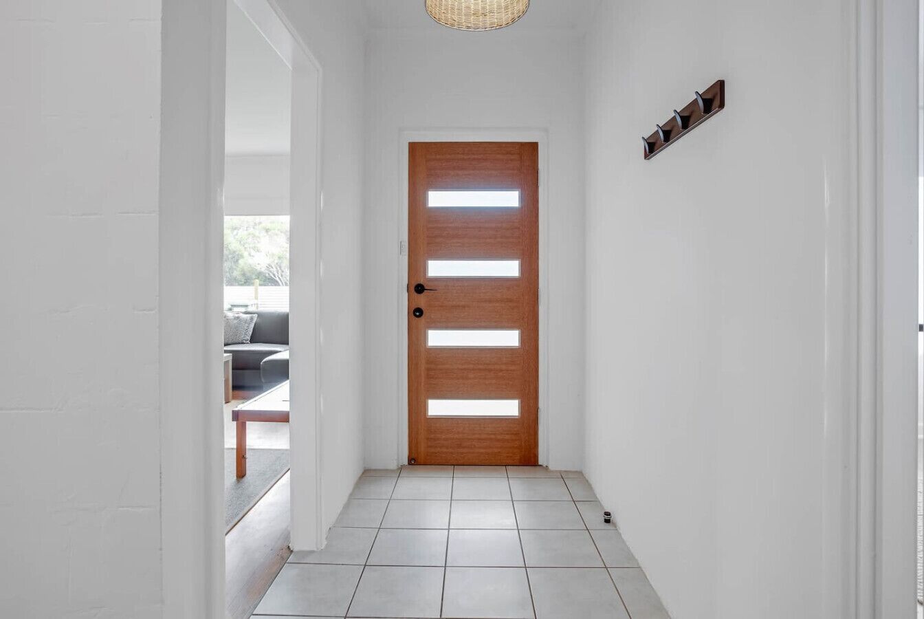 Un pasillo con puerta de madera, paredes blancas y suelo de baldosas.