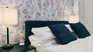 Ein ordentlich bezogenes Bett mit weißen und dunkelblauen Kissen, einem schwarzen Kopfteil und einem Nachttisch mit Lampe und Blumenvase.