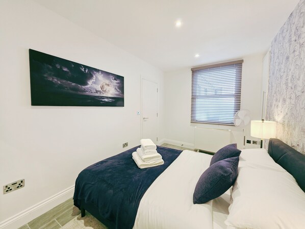 A bedroom with a bed, a night sky painting, a window with blinds, a standing fan, and a lamp.