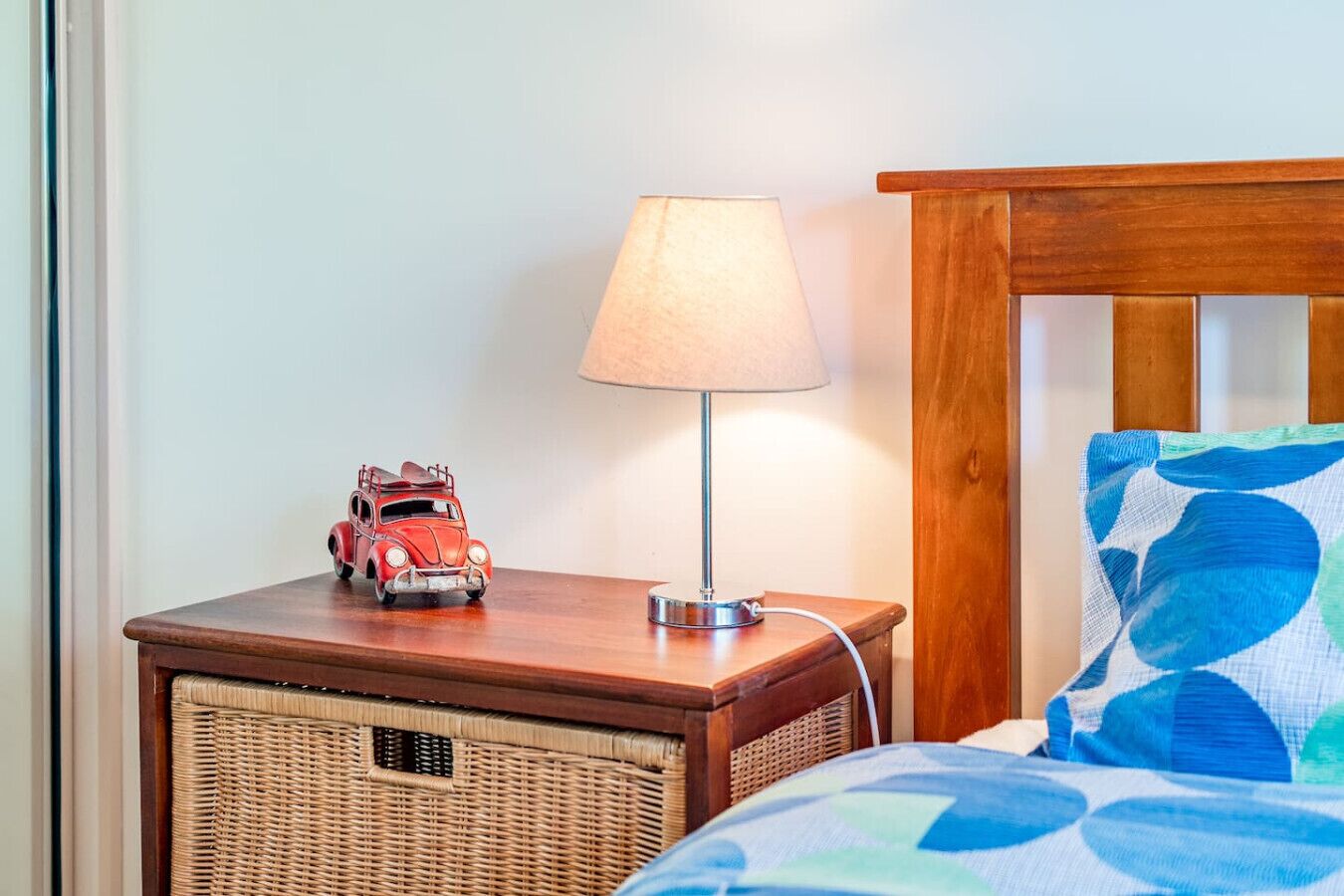 Una mesita de noche de madera con una lámpara, una cesta de mimbre, un coche de juguete y una cama con una almohada de estampado azul y verde.