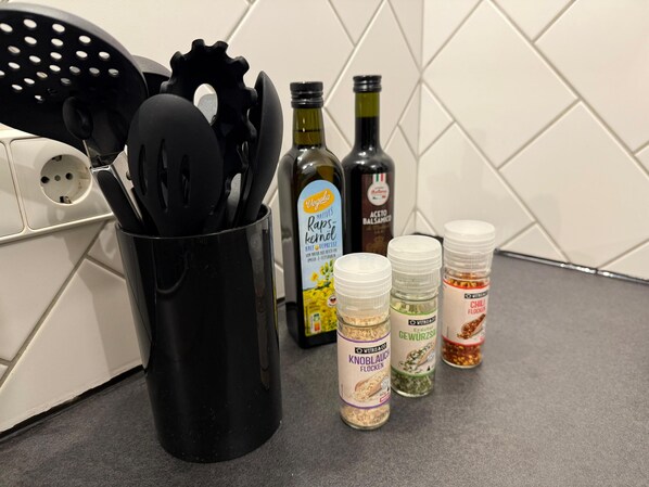 A kitchen counter with a black utensil holder, a bottle of olive oil, a bottle of balsamic vinegar, and three spice jars.