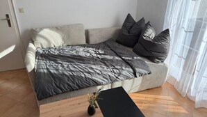 Une chambre à coucher moderne avec un lit, des oreillers, une petite table sur laquelle est posé un vase, et une fenêtre ornée de rideaux légers.