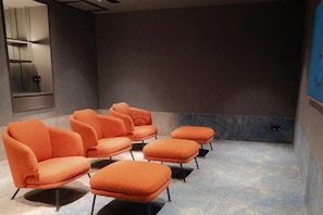 A room with a row of orange chairs and a large screen, likely for presentations or movies. - CapsuleTransit MAX - Terminal 2 (Sepang)