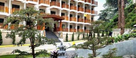 A multi-story hotel with balconies, a landscaped garden with trees and a pond, and a clear view of the building's entrance.