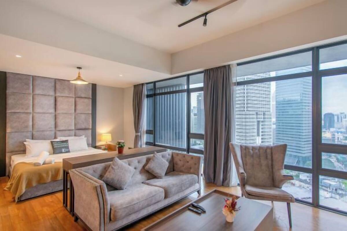 Una habitación de hotel moderna con cama, sofá, sillón, ventanas amplias y vistas a la ciudad.