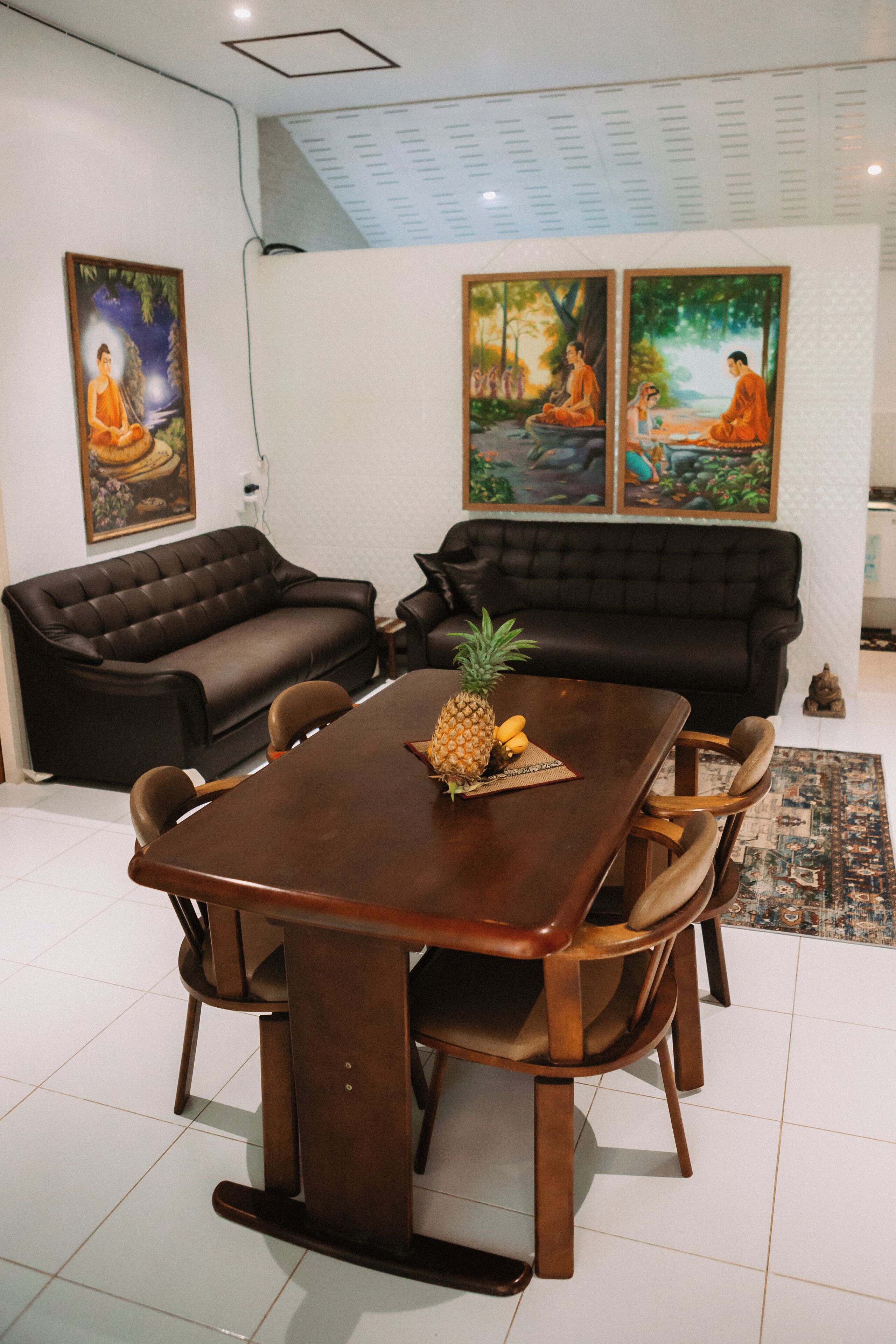 A dining area with a wooden table and chairs, a pineapple, and a Buddha statue. There are two black sofas and a patterned rug.
