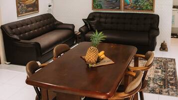 A dining area with a wooden table and chairs, a pineapple, and a Buddha statue. There are two black sofas and a patterned rug.