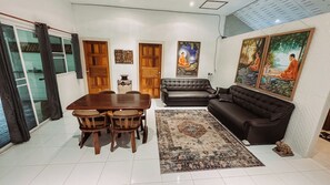 Una sala de estar con una mesa de comedor, sillas, un sofá y una alfombra. Hay dos puertas y una ventana con cortinas.
