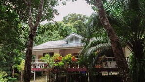 Una casa rodeada de densa vegetación tropical, con un porche cubierto y techo de tejas.