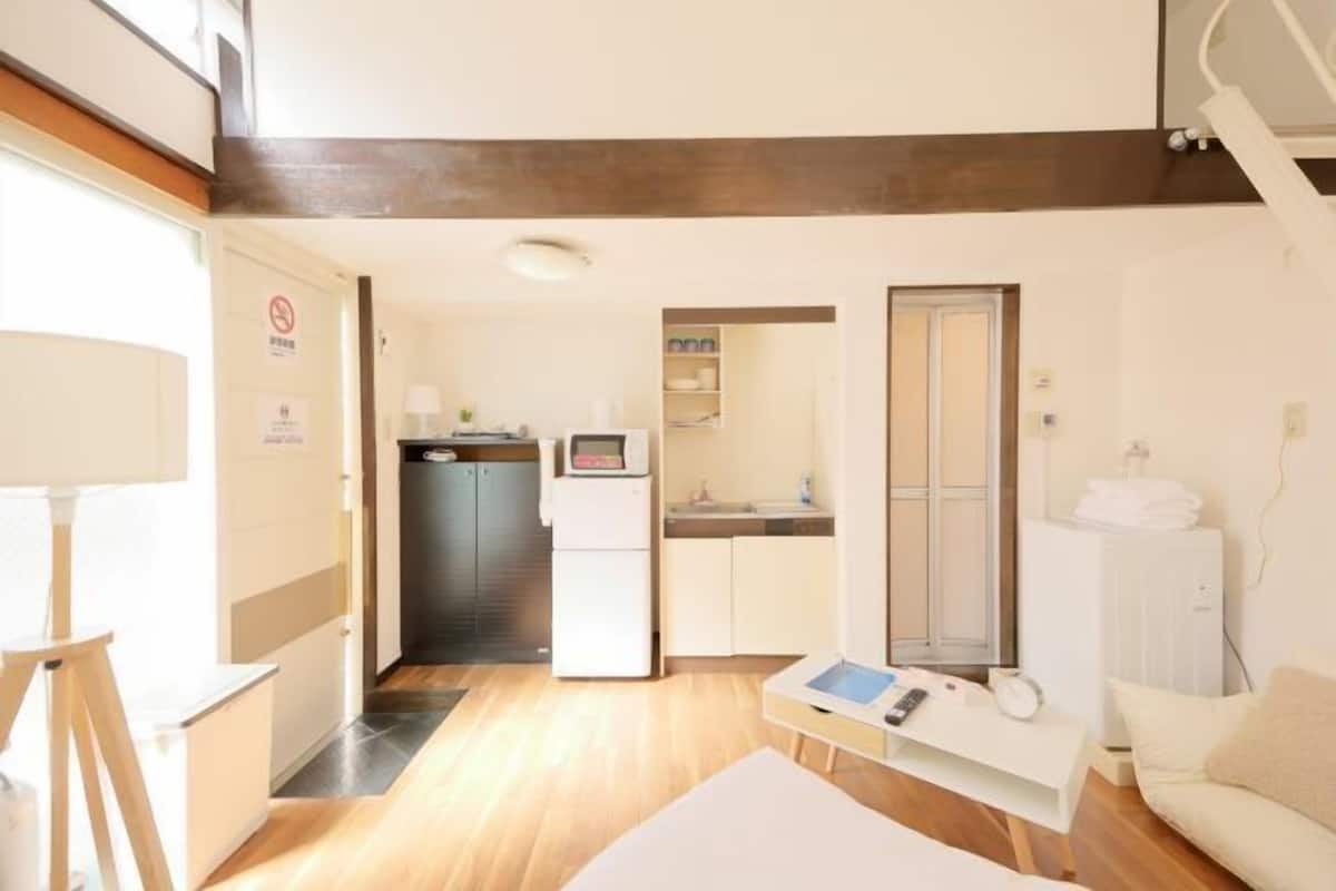 A modern hotel room with a wooden floor, a white sofa, a small table with a lamp, a TV, and a kitchenette in the background.