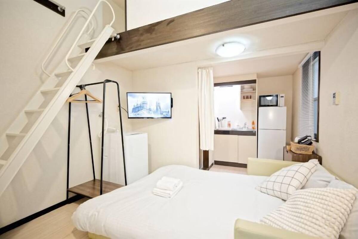 A bedroom with a bed, a TV mounted on the wall, a kitchen area visible through an open door, and a white staircase.