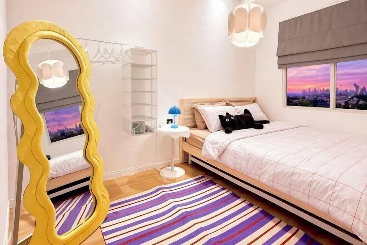 Een slaapkamer met een bed, een spiegel, een nachtkastje, een lamp, een raam met uitzicht op een stadsbeeld en een gestreept vloerkleed.