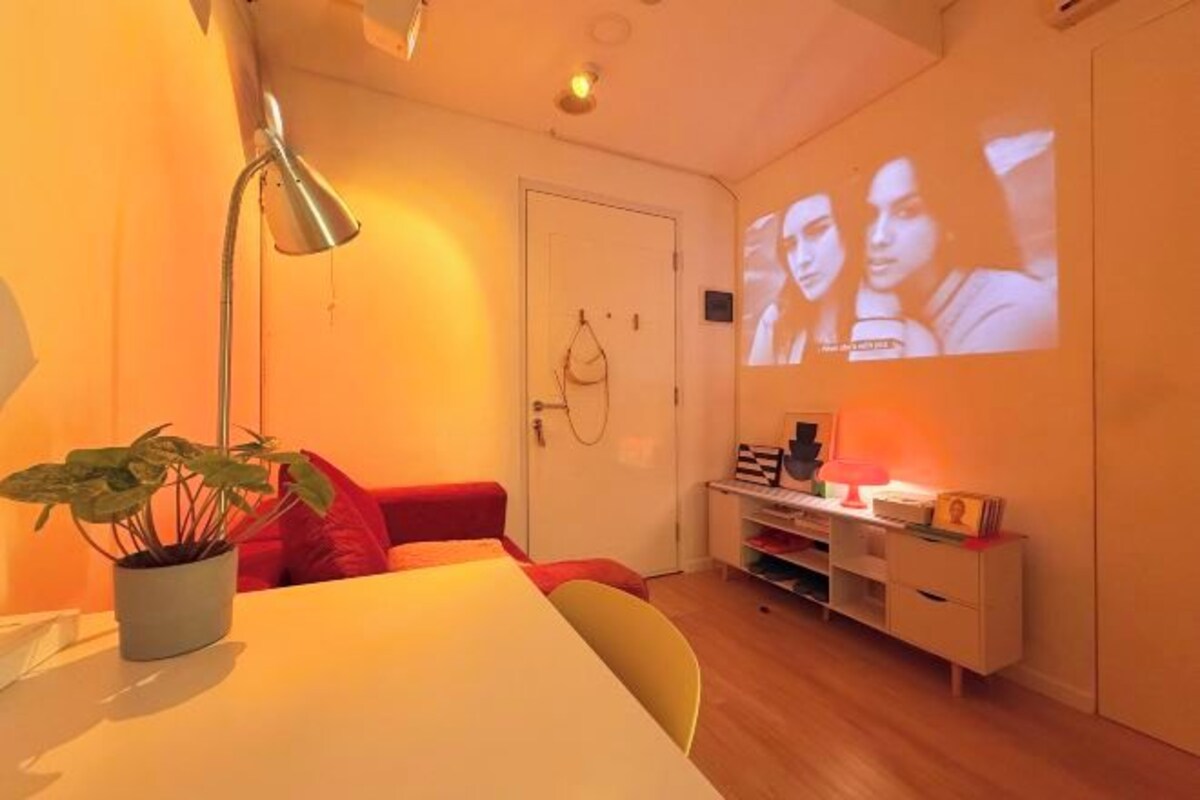 Ein Zimmer mit einem Bett, einem Schreibtisch, einem Sessel, einer Pflanze, einer Lampe und einer Projektionsleinwand.