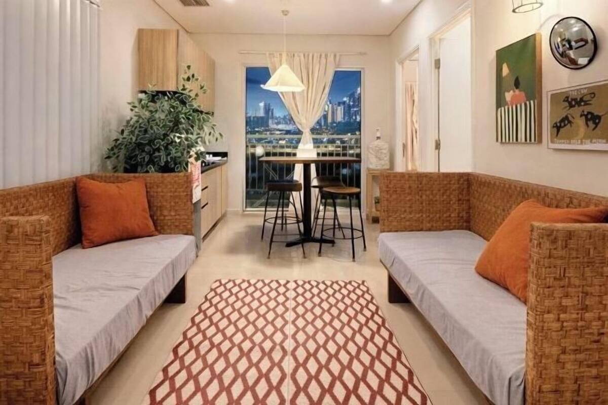 Een kamer met twee rieten banken, een kamerbreed tapijt met patroon, een eettafel met stoelen en een uitzicht op een stadsbeeld door een raam.