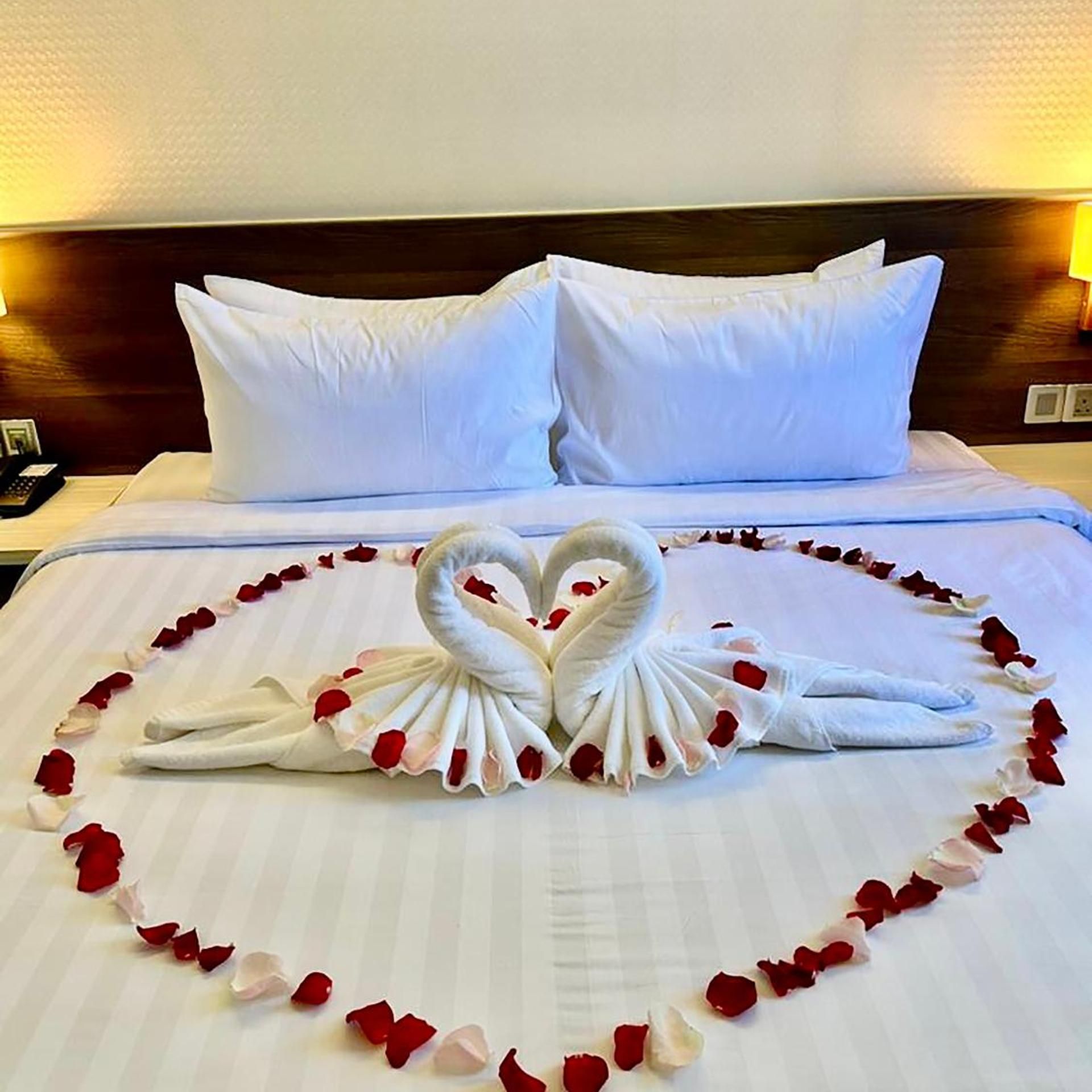 A bed with white linens, two pillows, and a heart-shaped towel arrangement made with towels and rose petals.