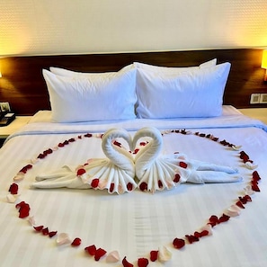 A bed with white linens, two pillows, and a heart-shaped towel arrangement made with towels and rose petals.