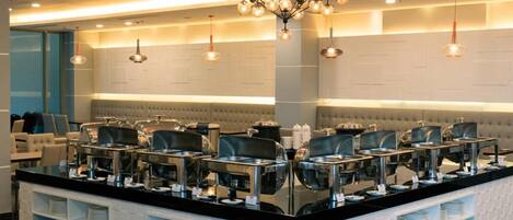 Un moderno vestíbulo de hotel con una isla central que ofrece servicio tipo buffet, áreas de estar y un candelabro.
