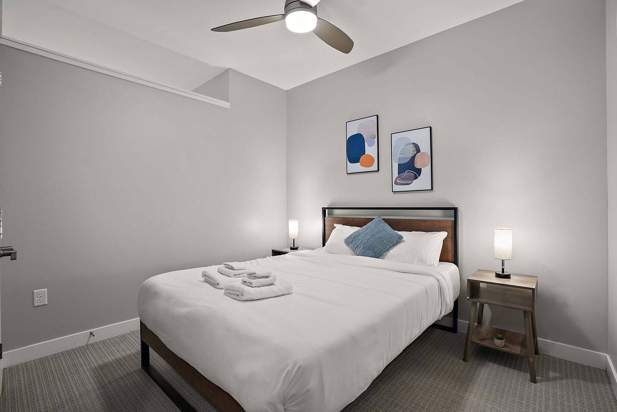 A neatly made bed with white linens and towels, a ceiling fan, two wall-mounted artworks, a bedside table with a lamp, and a small potted plant.
