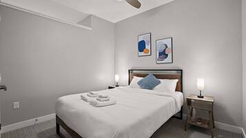 A neatly made bed with white linens and towels, a ceiling fan, two wall-mounted artworks, a bedside table with a lamp, and a small potted plant.