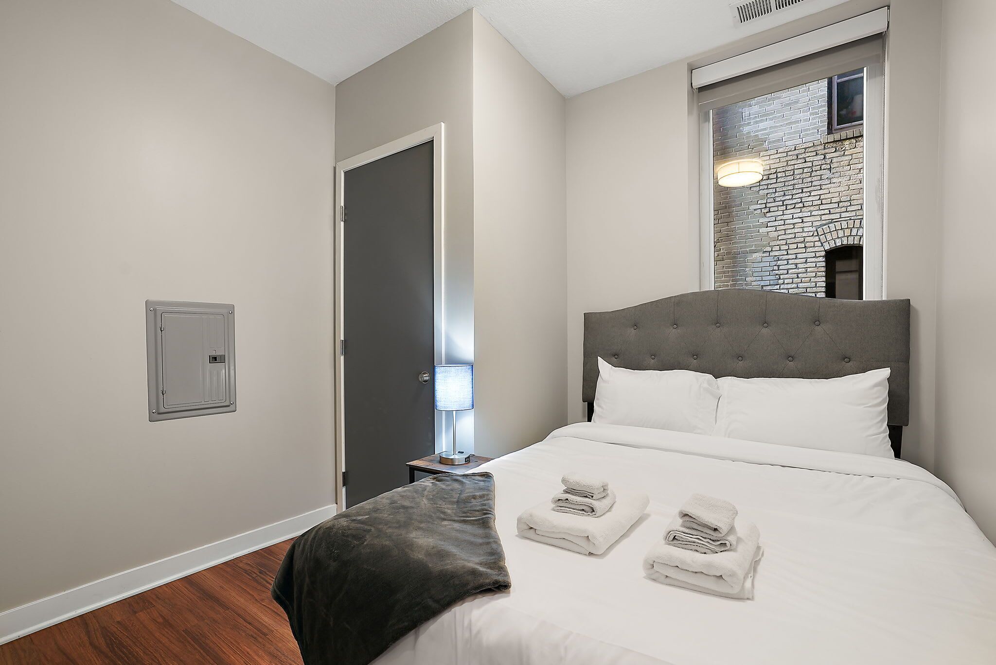 Ett hotellrum med en stor säng, en grå sänggavel och ett fönster med utsikt över en tegelvägg.