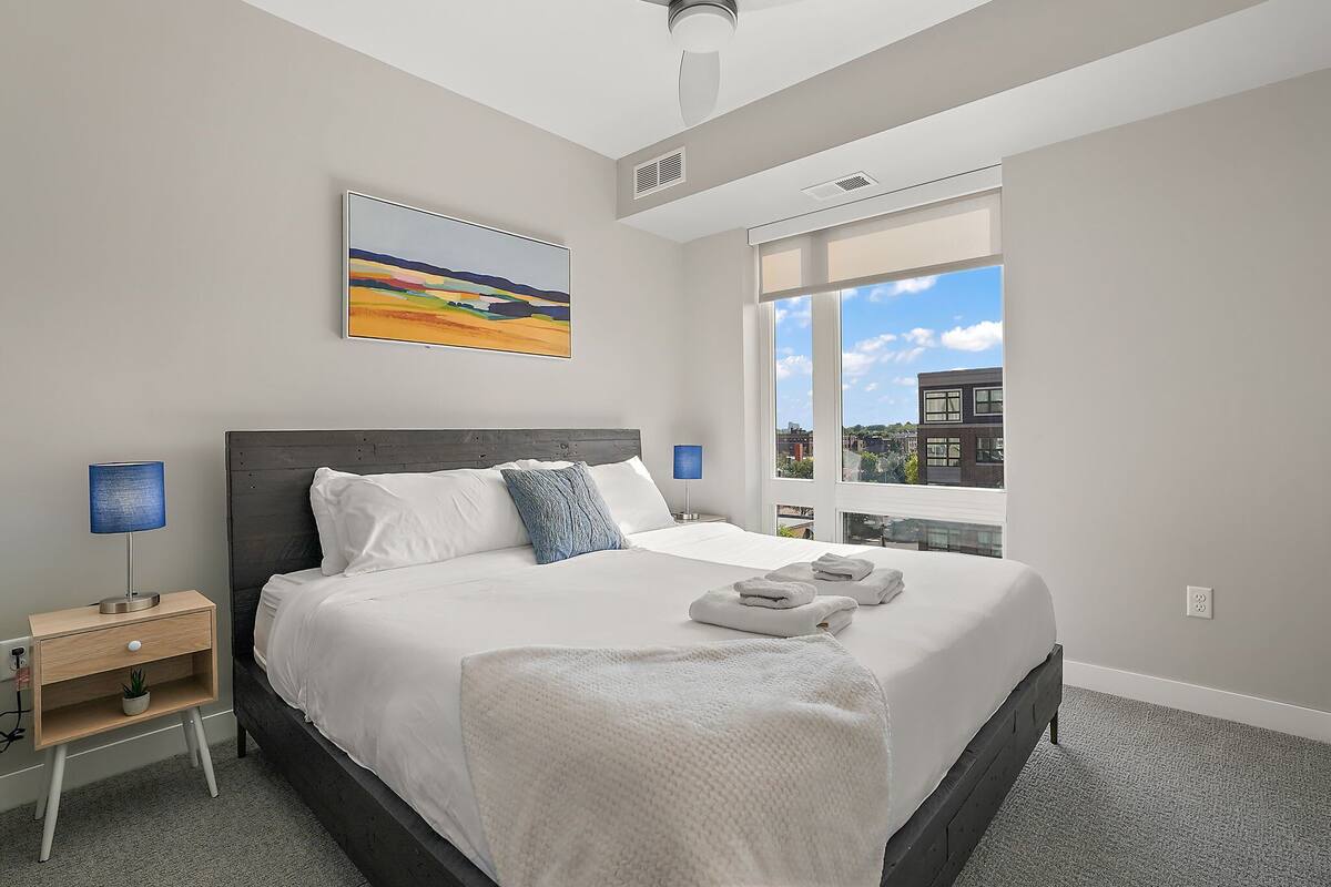 Un dormitorio moderno con una cama grande, una mesita de noche, una lámpara de pared y una ventana con vistas a la ciudad.