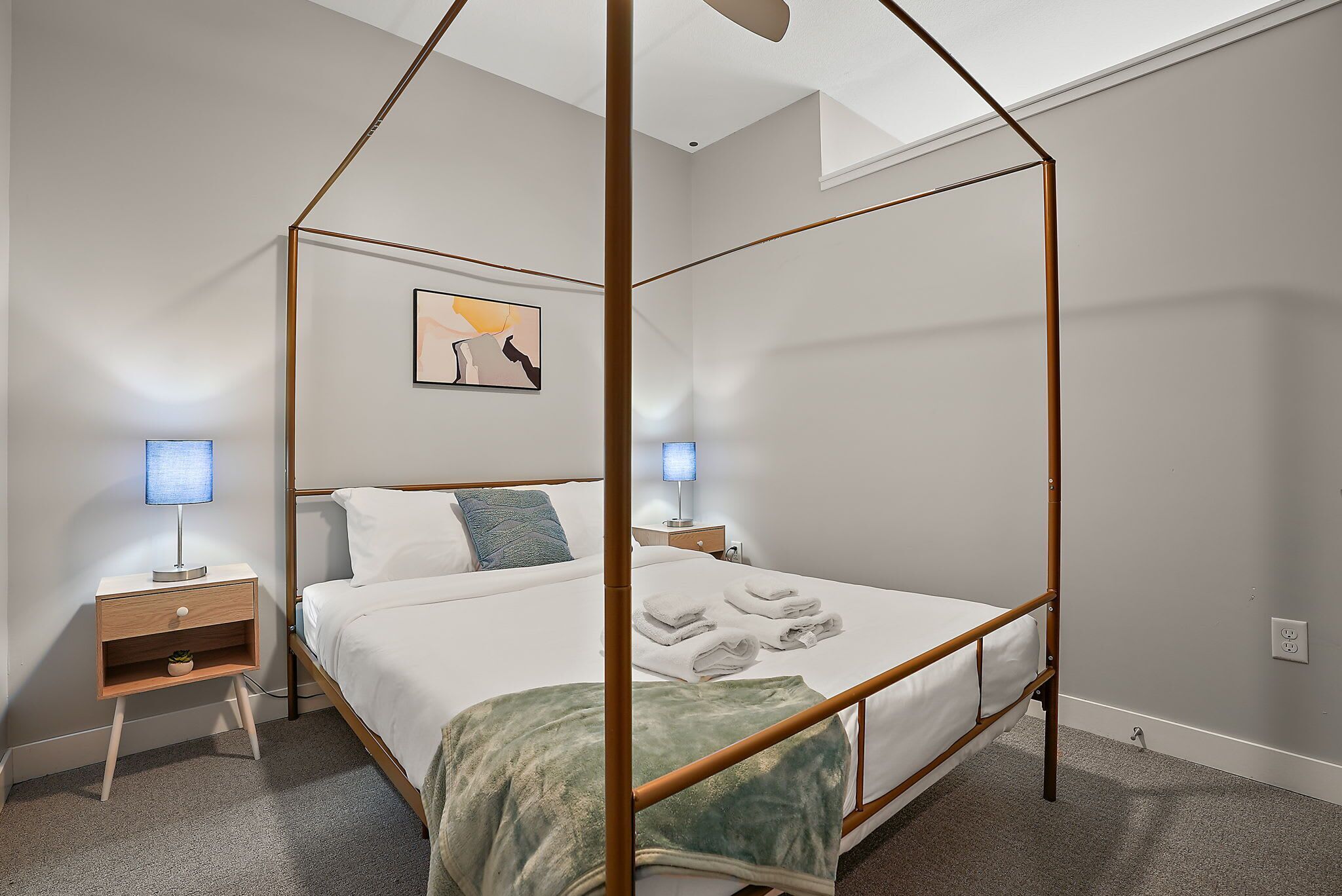 A neatly arranged bedroom with a four-poster bed, bedside tables with lamps, a small shelf, and a wall-mounted artwork.