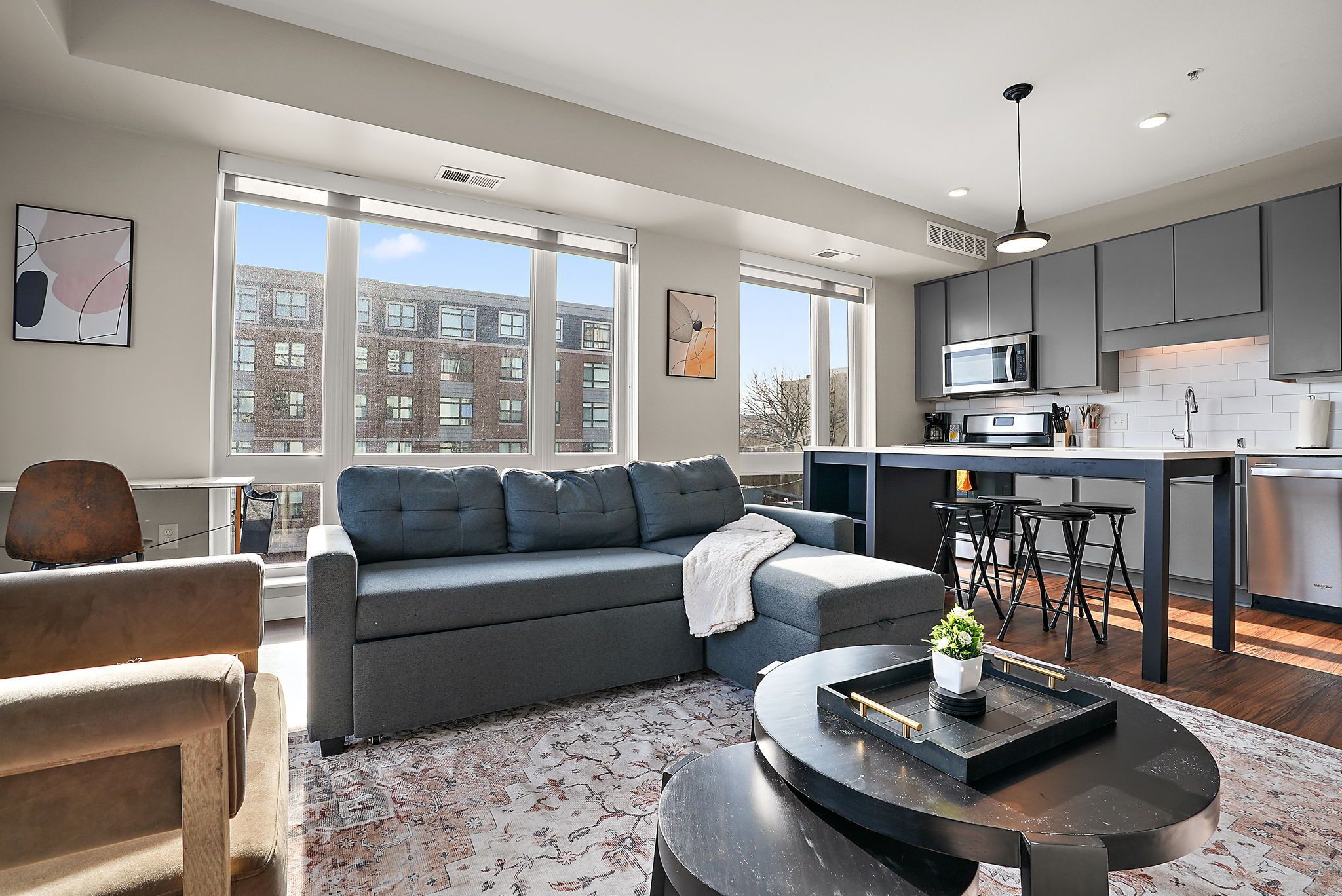A modern living room with a grey sectional sofa, a coffee table with a plant, and a view of a cityscape through large windows.